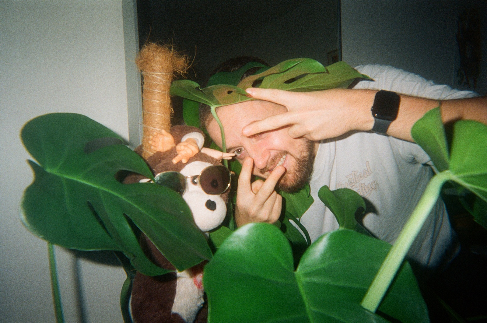 Jimmy Doré peeking through monstera leaves, making a peace sign next to a stuffed monkey